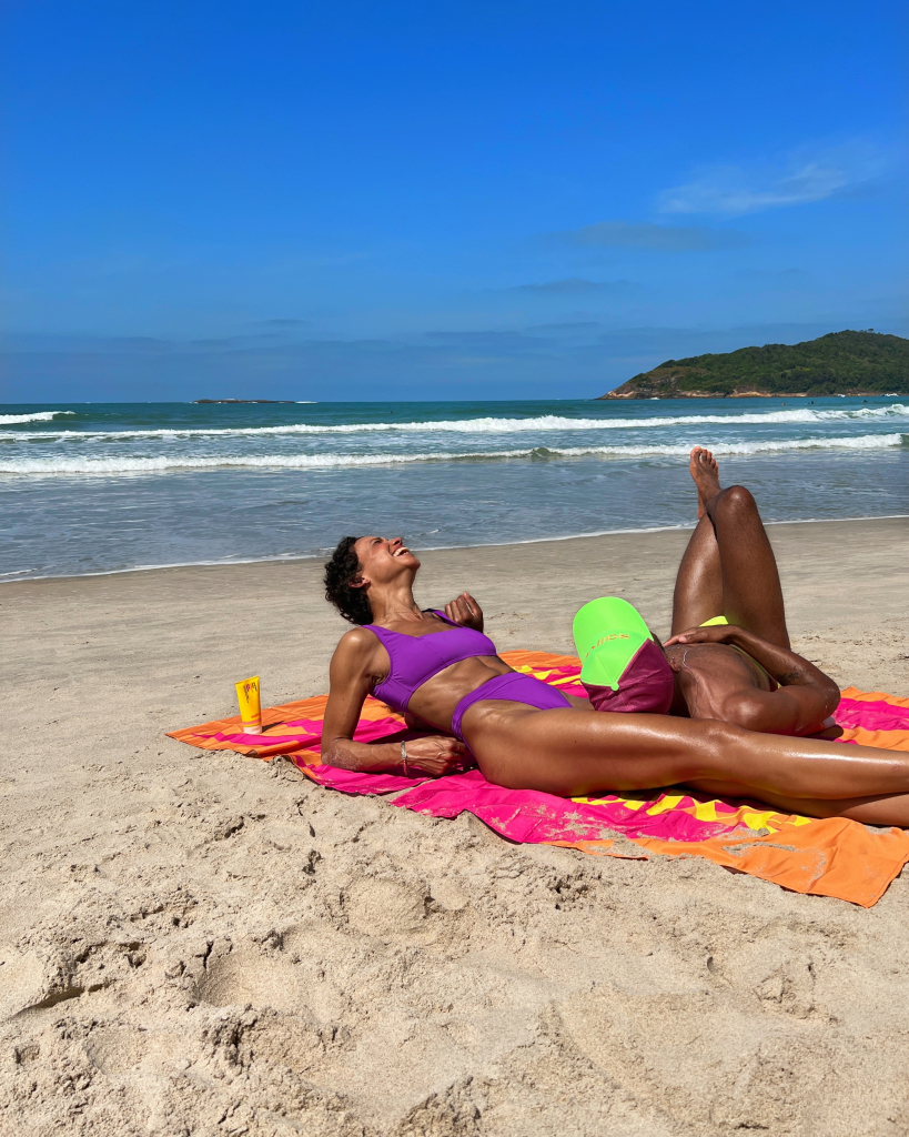mulher vestindo biquíni roxo, deitada em uma canga na praia, com o logo da sallve estampado. homem deitado na perna esquerda da mulher, usando um boné verde com o logo da sallve grafado.