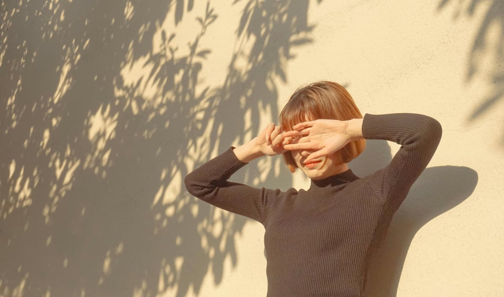 imagem mostra uma mulher branca, de cabelos curtos avermelhados, que protege o rosto do sol com as mãos. ela está encostada em um muro branco, que tem sombras de uma árvore. o sol é um dos principais fatores de piora do melasma